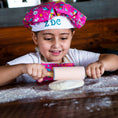 Load image into Gallery viewer, Child in a colorful chef's hat rolling out dough with a pink rolling pin on a wooden table.