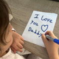Load image into Gallery viewer, Child writing 'I Love You Daddy' on a piece of paper with a blue marker.