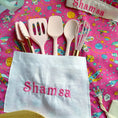 Load image into Gallery viewer, Kitchen utensils including spatulas and a whisk on a colorful pink background with 'Shamsa' embroidered on a towel.