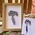 Load image into Gallery viewer, Framed paper cutout of a stylized face next to a child's drawing on a wooden surface.