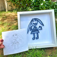 Load image into Gallery viewer, Framed paper cutout of a stick figure with a dress, next to a hand-drawn version outdoors.