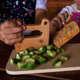 Load image into Gallery viewer, Children's hands using wooden kitchen tools to chop cucumbers on a cutting board.