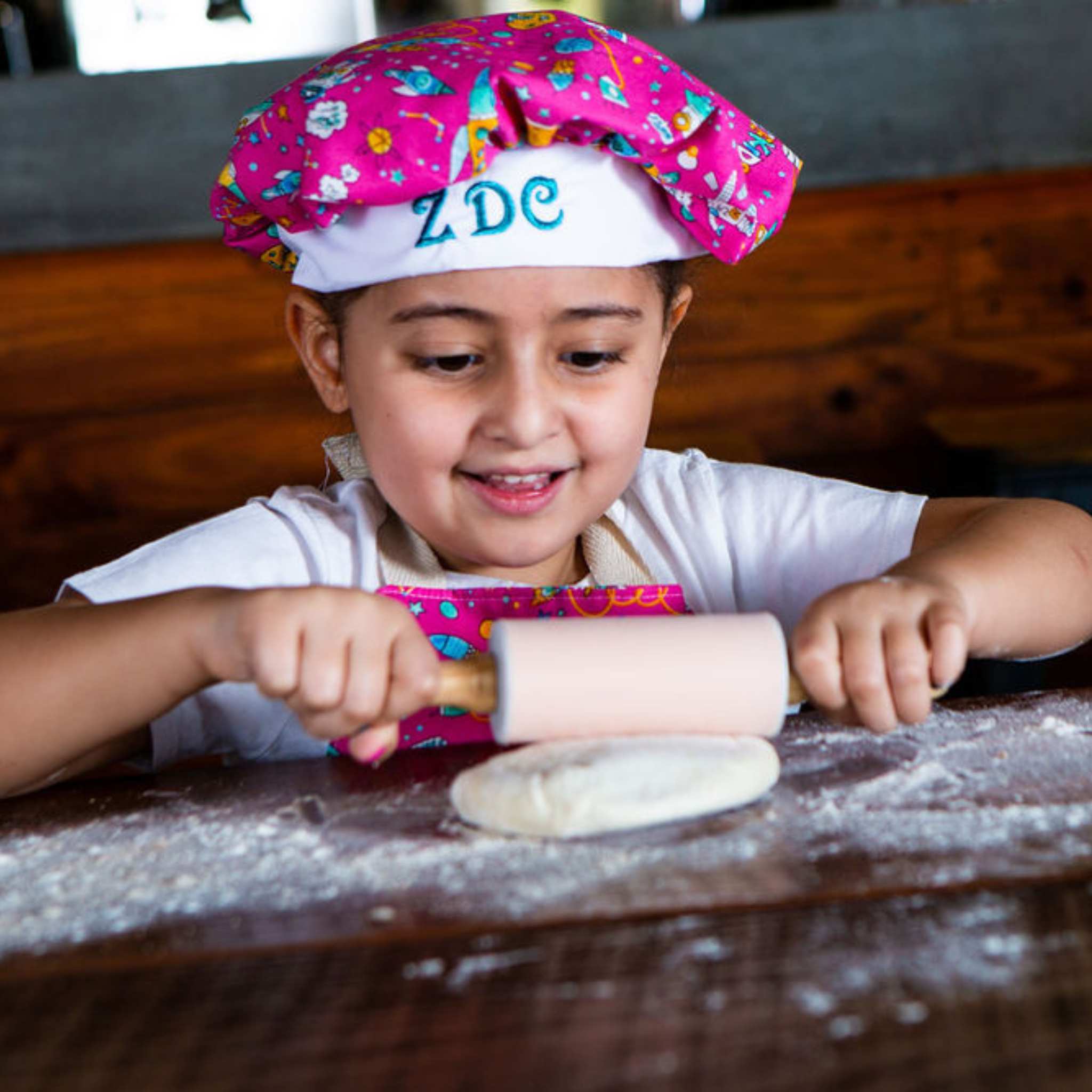 Child in a colorful chef's hat rolling out dough with a pink rolling pin on a wooden table.
