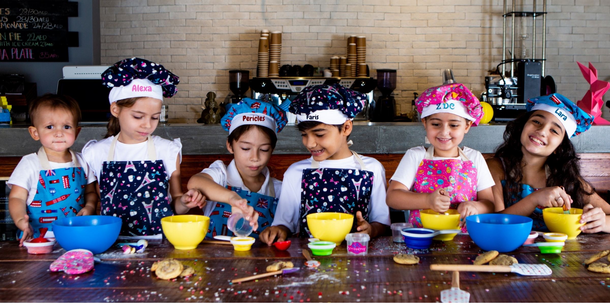 Children in a kitchen wearing colorful aprons and hats, engaged in cooking activities.