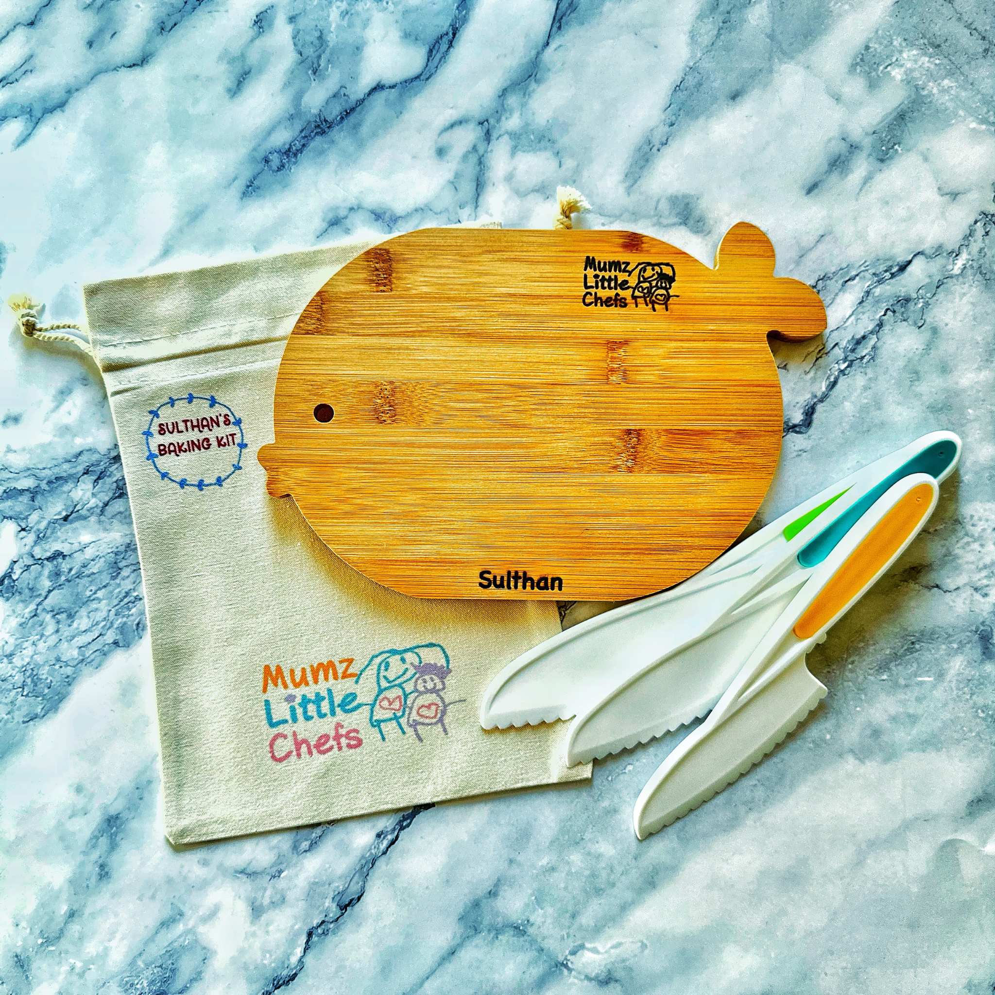 Wooden cutting board with 'Mumz Little Chefs' branding, accompanied by a knife and a branded bag on a marble surface.