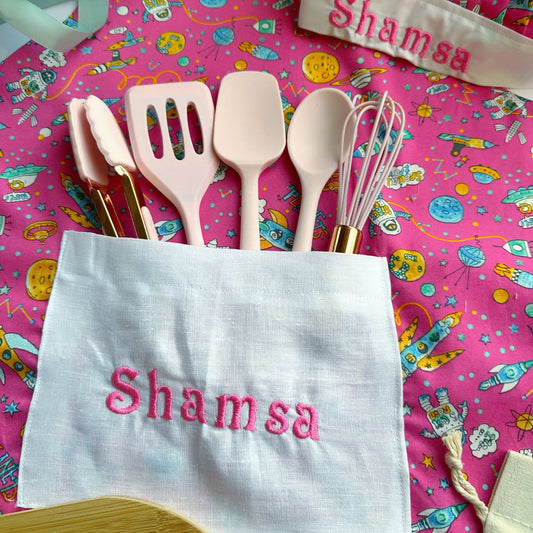 Kitchen utensils including spatulas and a whisk on a colorful pink background with 'Shamsa' embroidered on a towel.