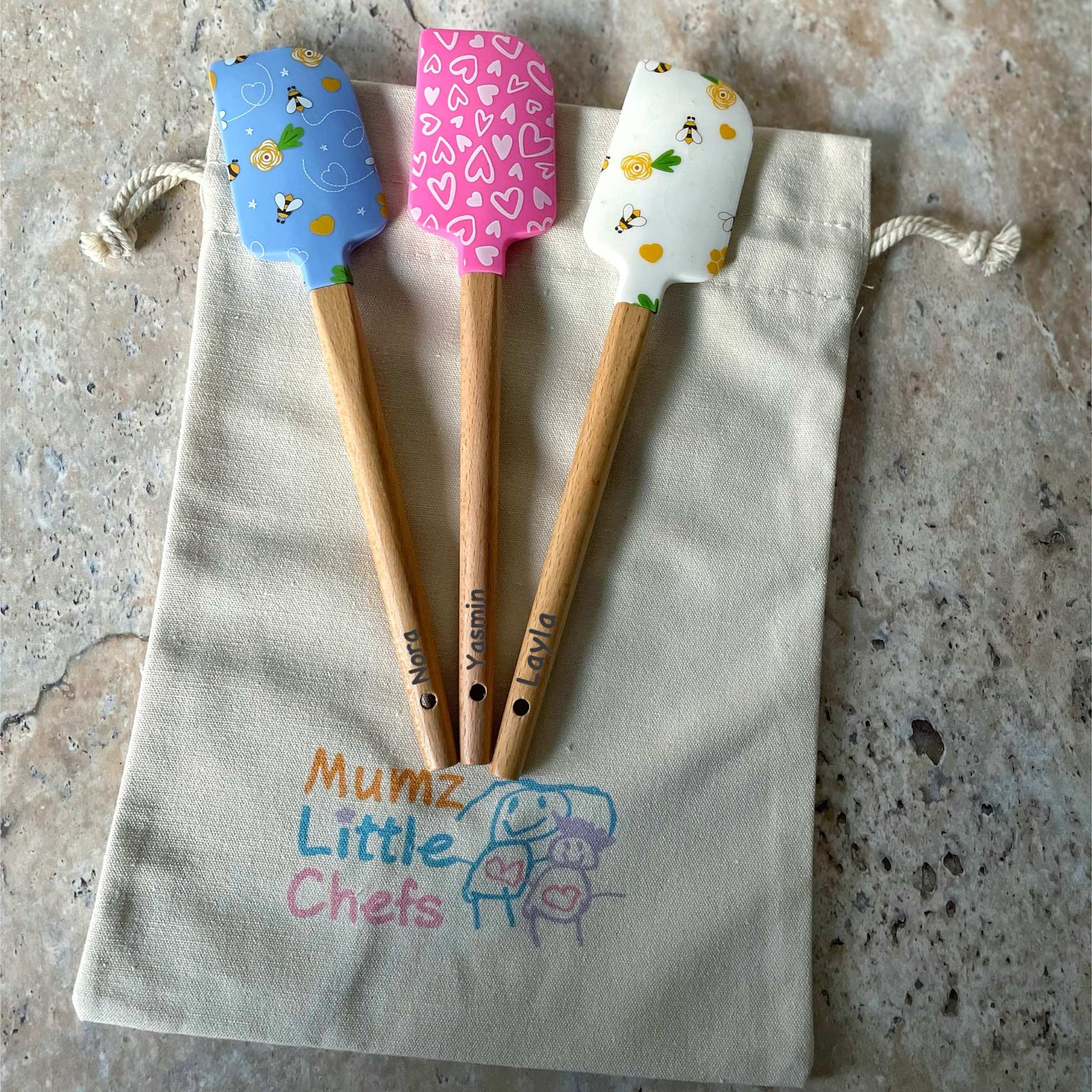 Three wooden spatulas with colorful handles on a 'Mumz Little Chefs' bag.