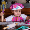 Load image into Gallery viewer, Child in a colorful chef's hat and apron holding a whisk, sitting at a table with a rolling pin and other kitchen tools.
