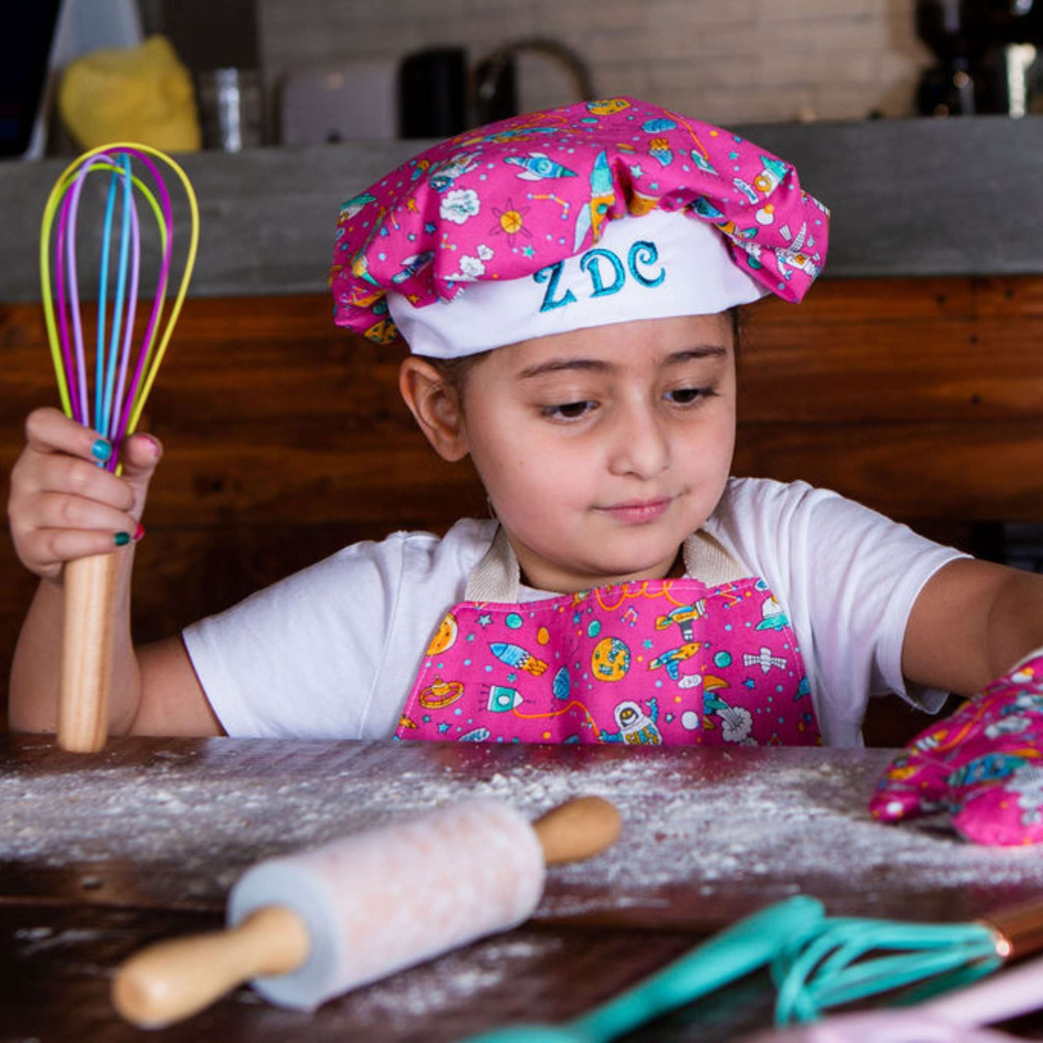 Child in a colorful chef's hat and apron holding a whisk, sitting at a table with a rolling pin and other kitchen tools.