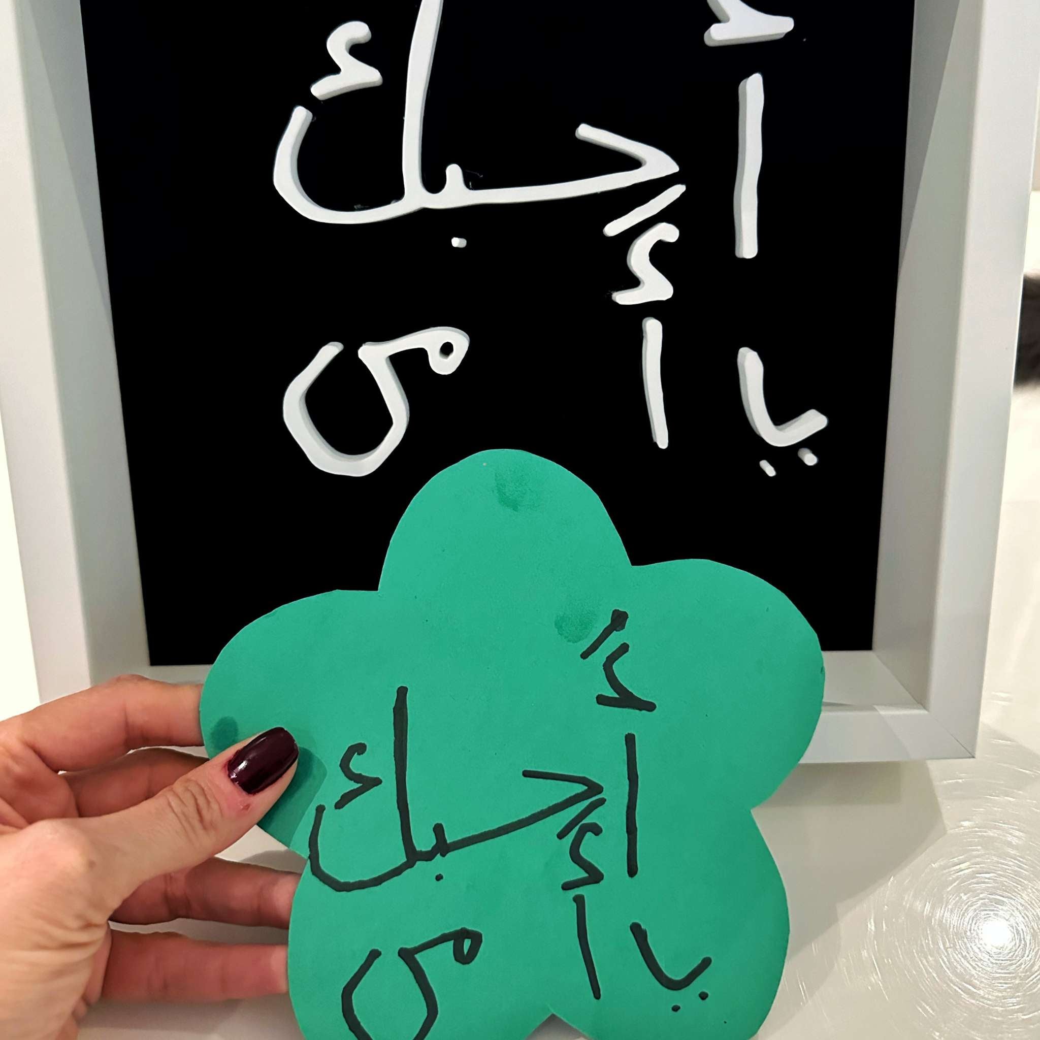 Hand holding a green flower-shaped card with Arabic text in front of a blackboard with similar text.