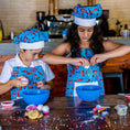 Load image into Gallery viewer, Two children in a kitchen wearing blue aprons and hats, engaged in a cooking activity.
