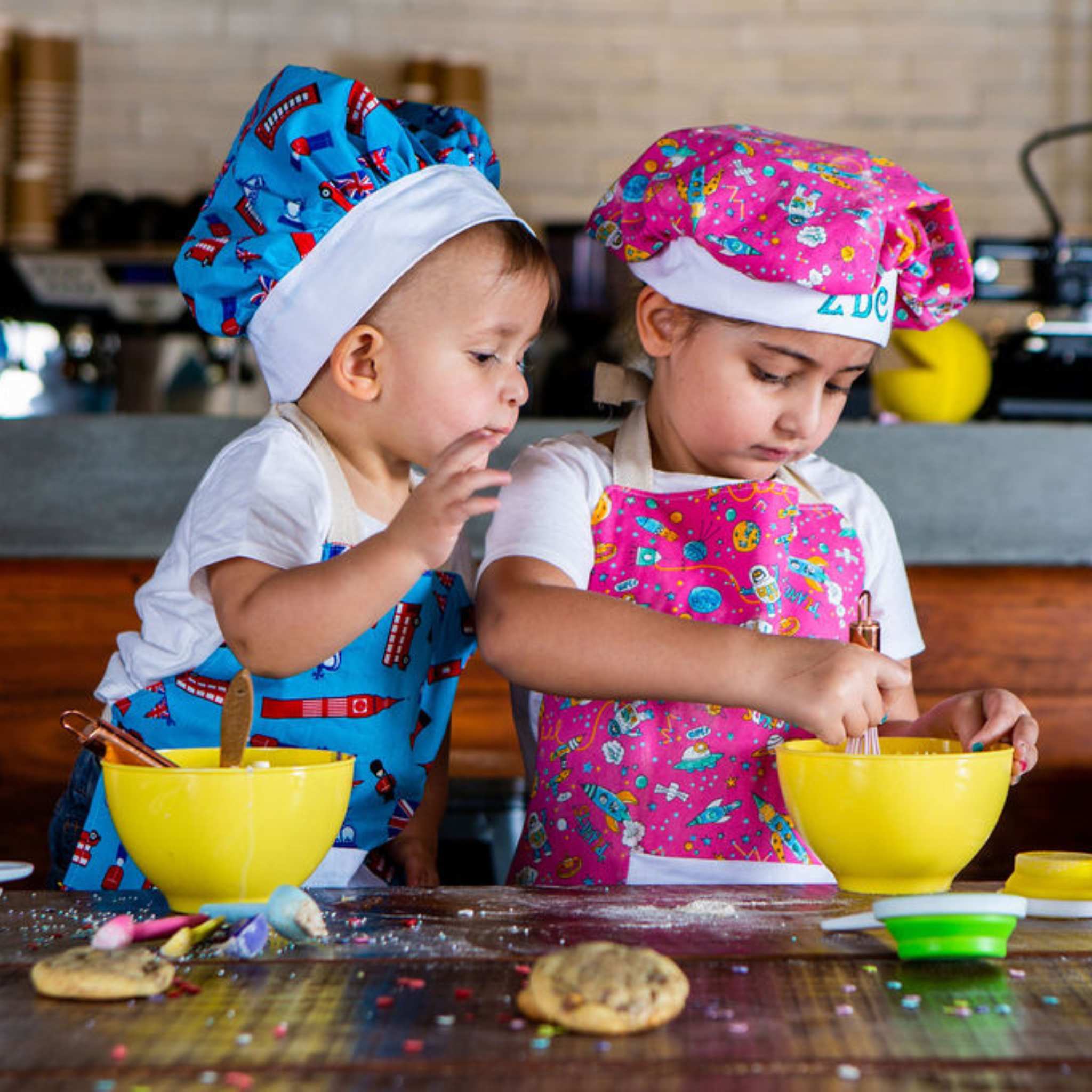 Two children in colorful aprons and chef hats mixing ingredients in yellow bowls.
