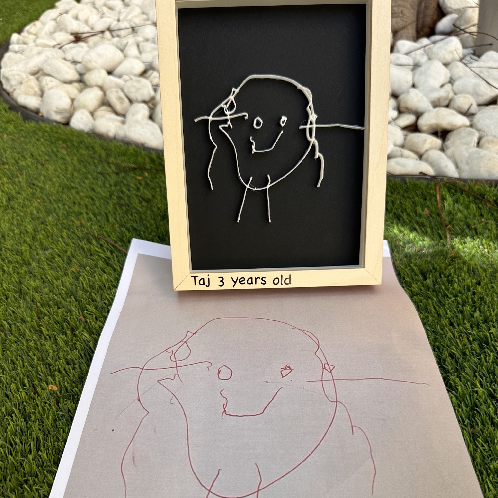 Child's drawing of a person on a blackboard with 'Taj 3 years old' written below, placed on grass.