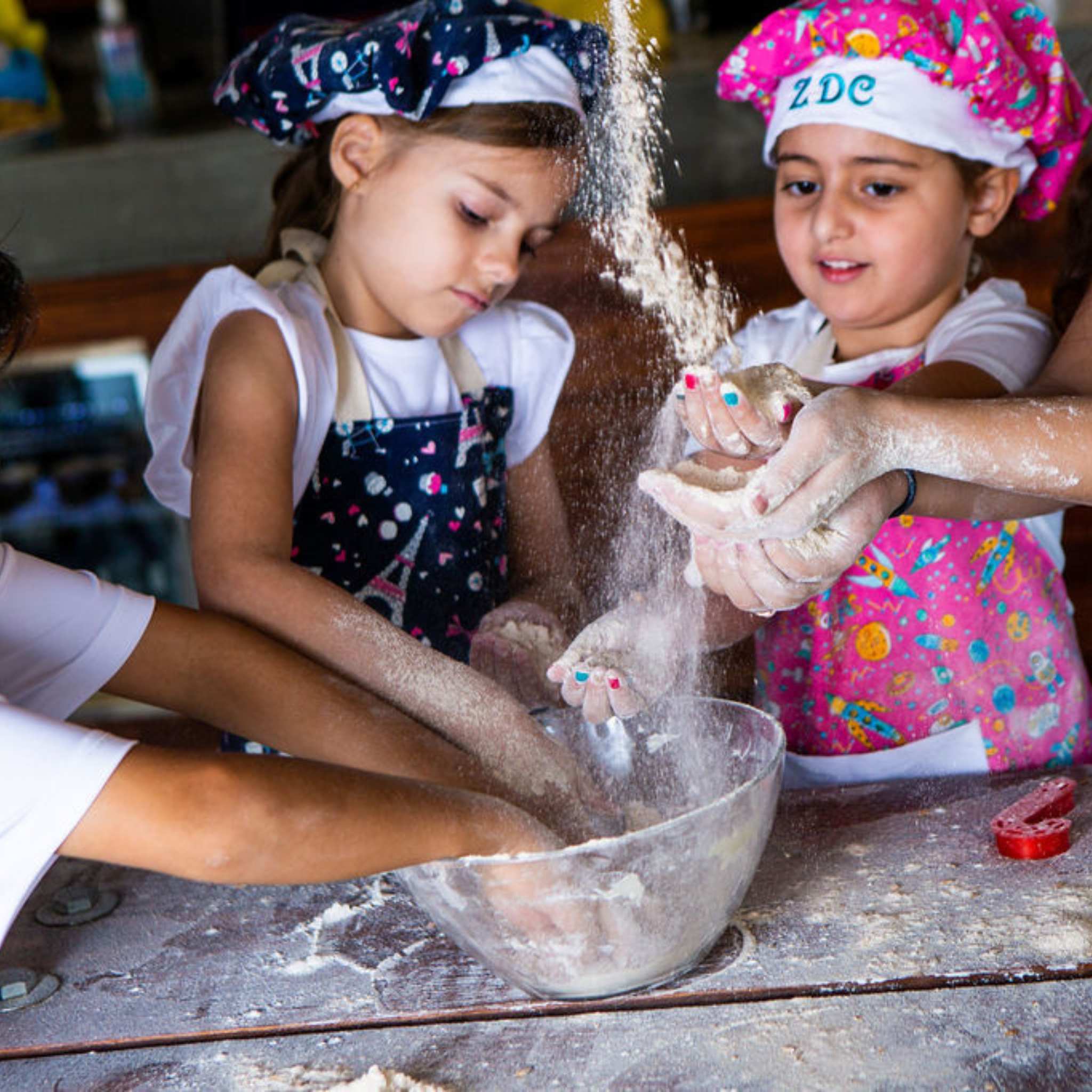 Two young girls in chef hats and aprons mixing flour in a bowl.