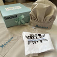 Load image into Gallery viewer, Chef's hat, apron, and towel with 'Mansour' branding on a kitchen counter.

