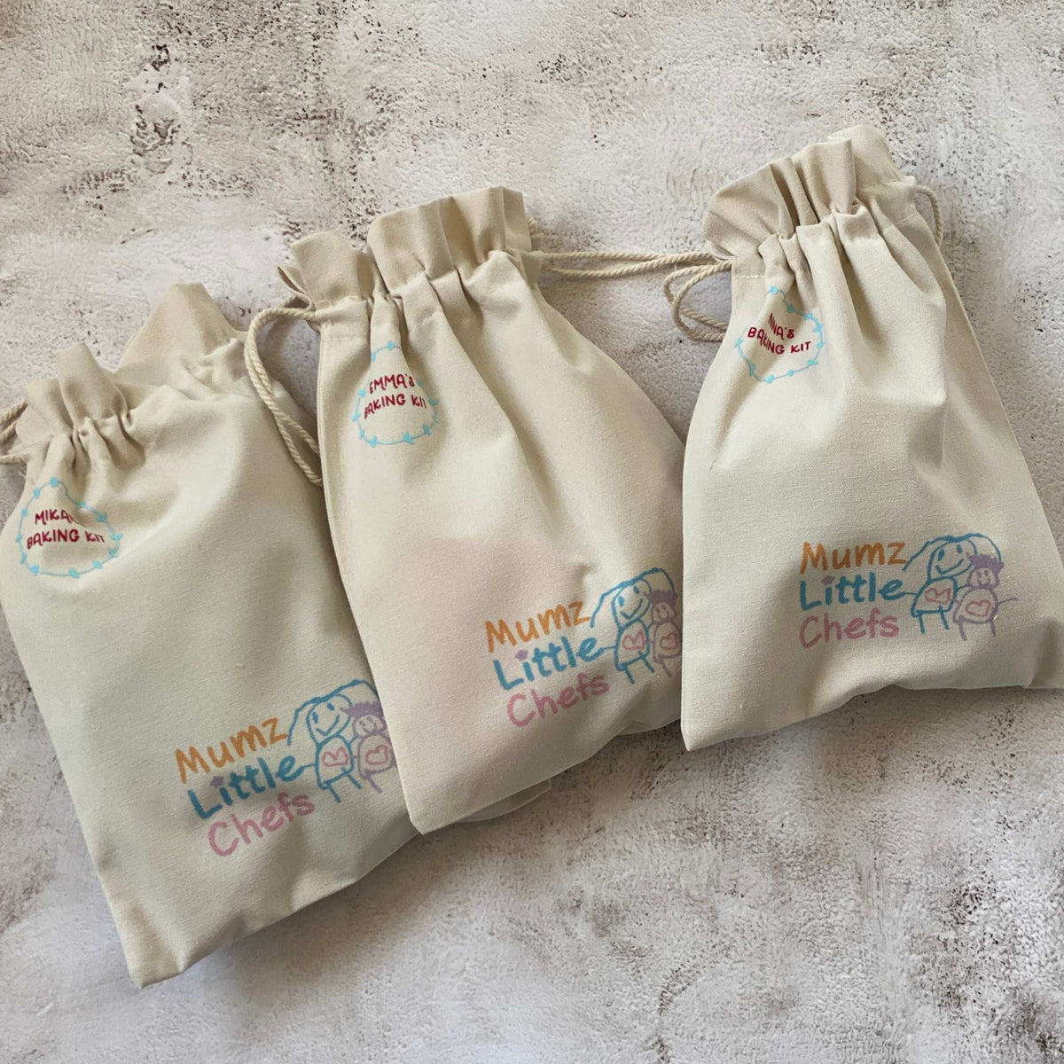 Three beige drawstring bags with 'Mumz Little Chefs' branding on a textured surface.