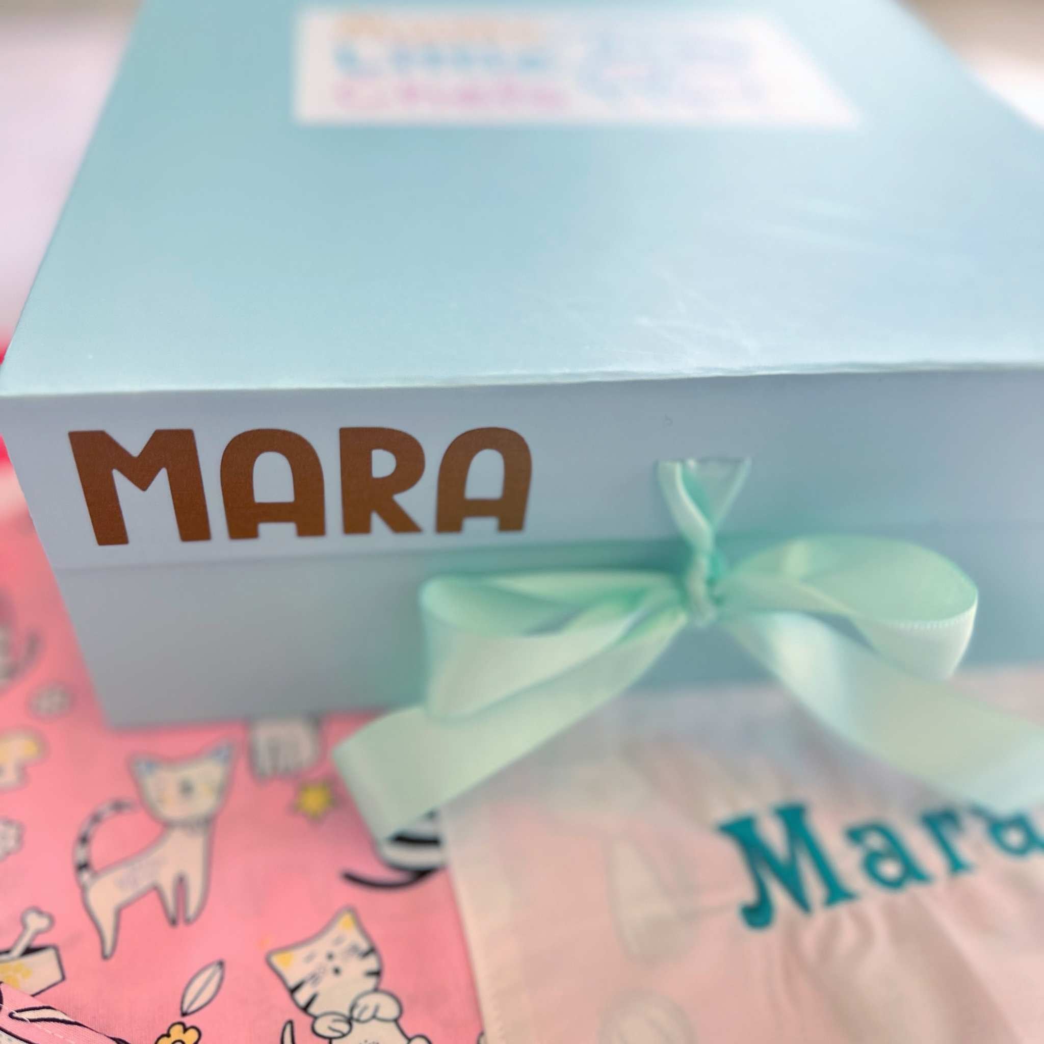 Gift box with a green ribbon labeled 'Mara' on a pink background with cat illustrations.