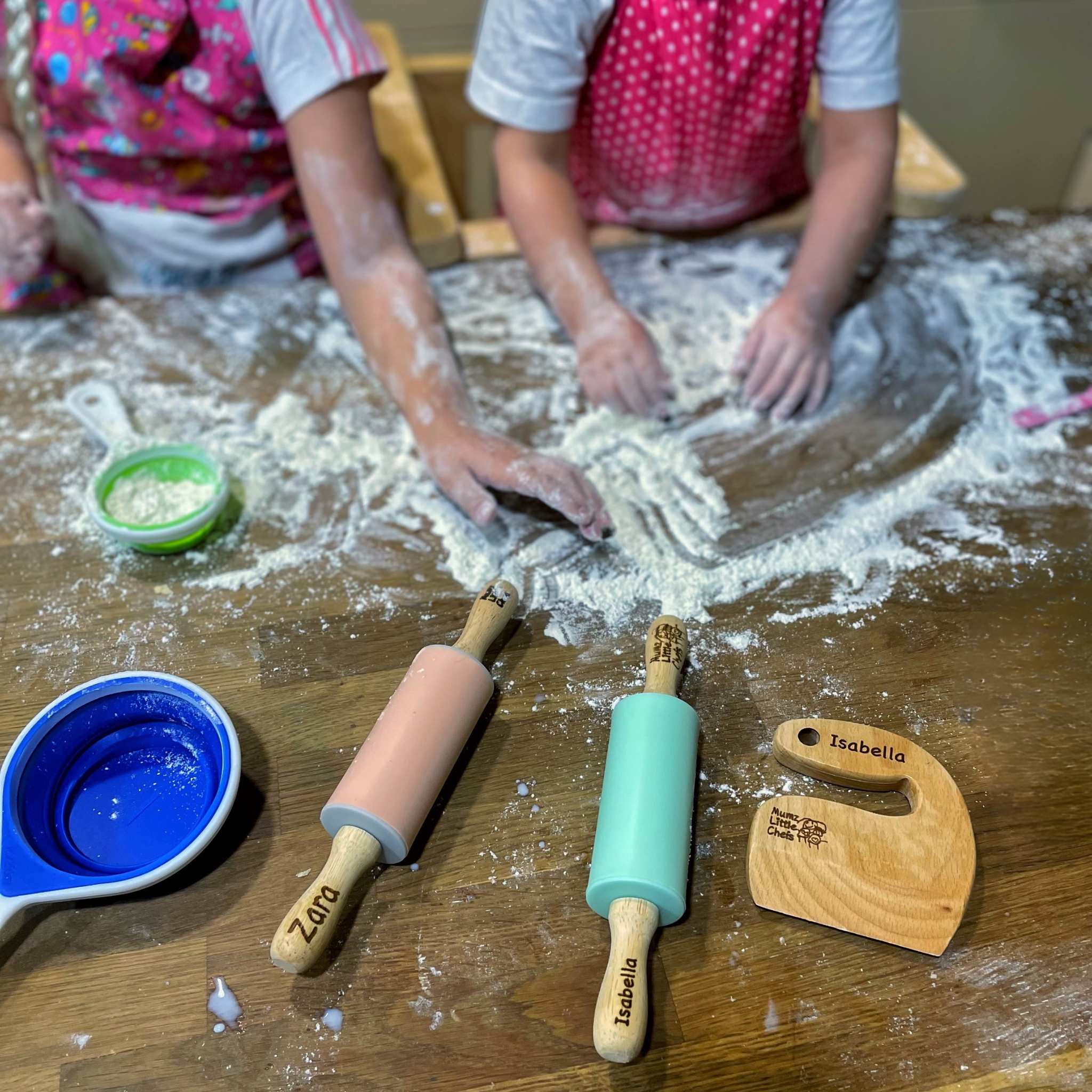 Children playing with flour on a wooden table with personalized kitchen tools.
