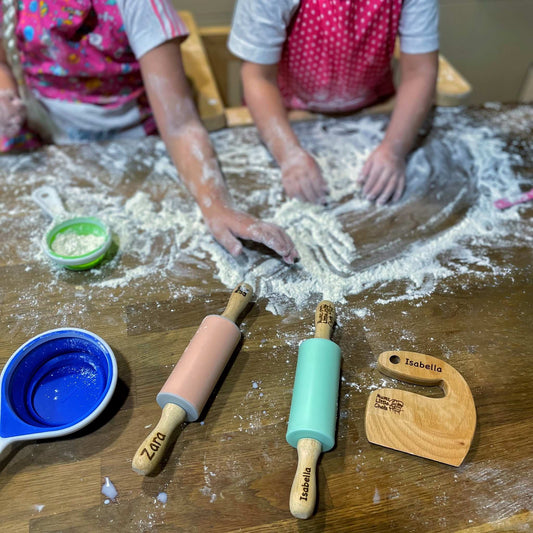 Children playing with flour on a wooden table with personalized kitchen tools.