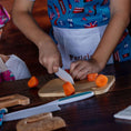 Load image into Gallery viewer, Child in a colorful apron cutting carrots on a wooden board with supervision.
