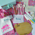 Load image into Gallery viewer, Kitchen set with pink apron, utensils, and cutting board on a colorful background with 'Hessa' branding.
