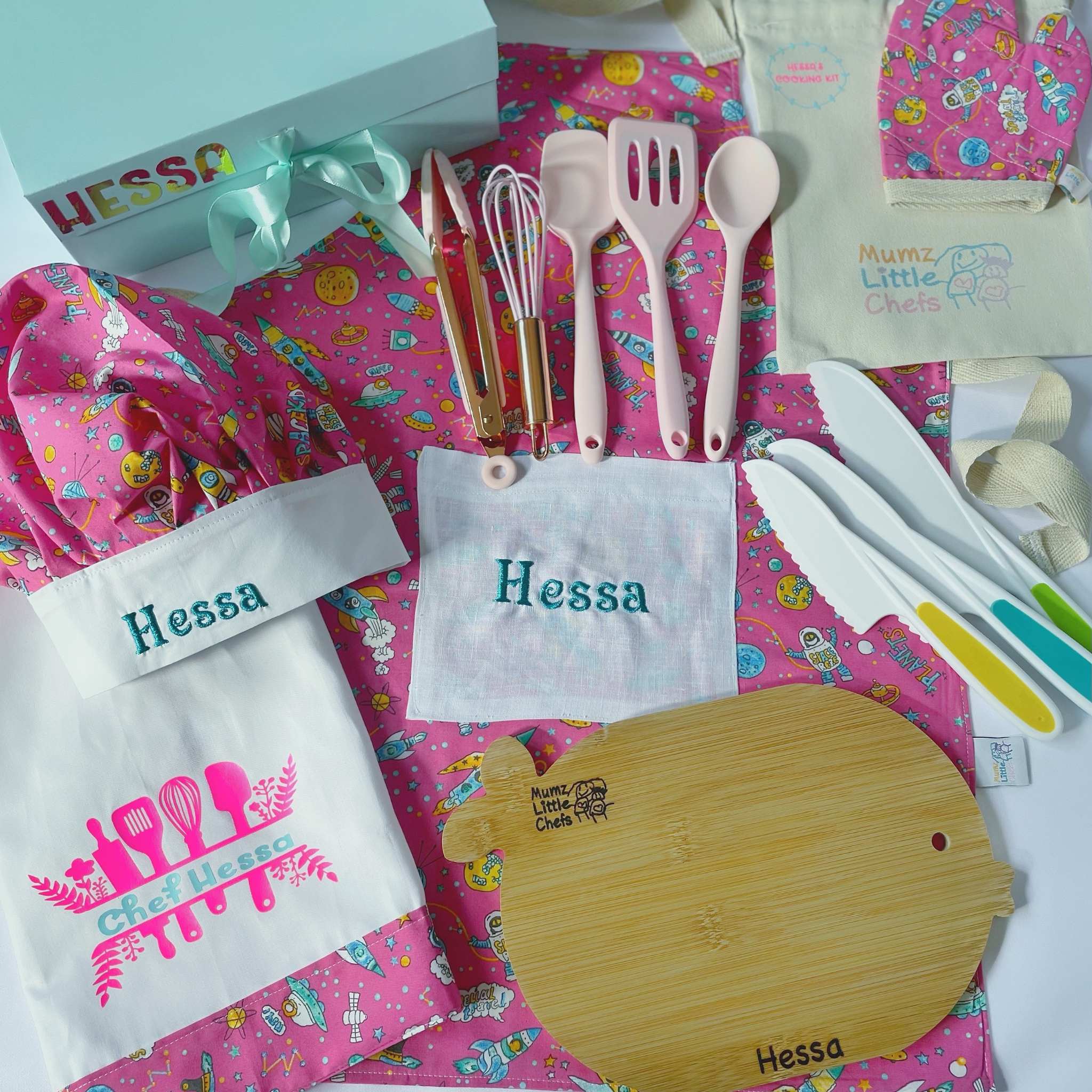 Kitchen set with pink apron, utensils, and cutting board on a colorful background with 'Hessa' branding.