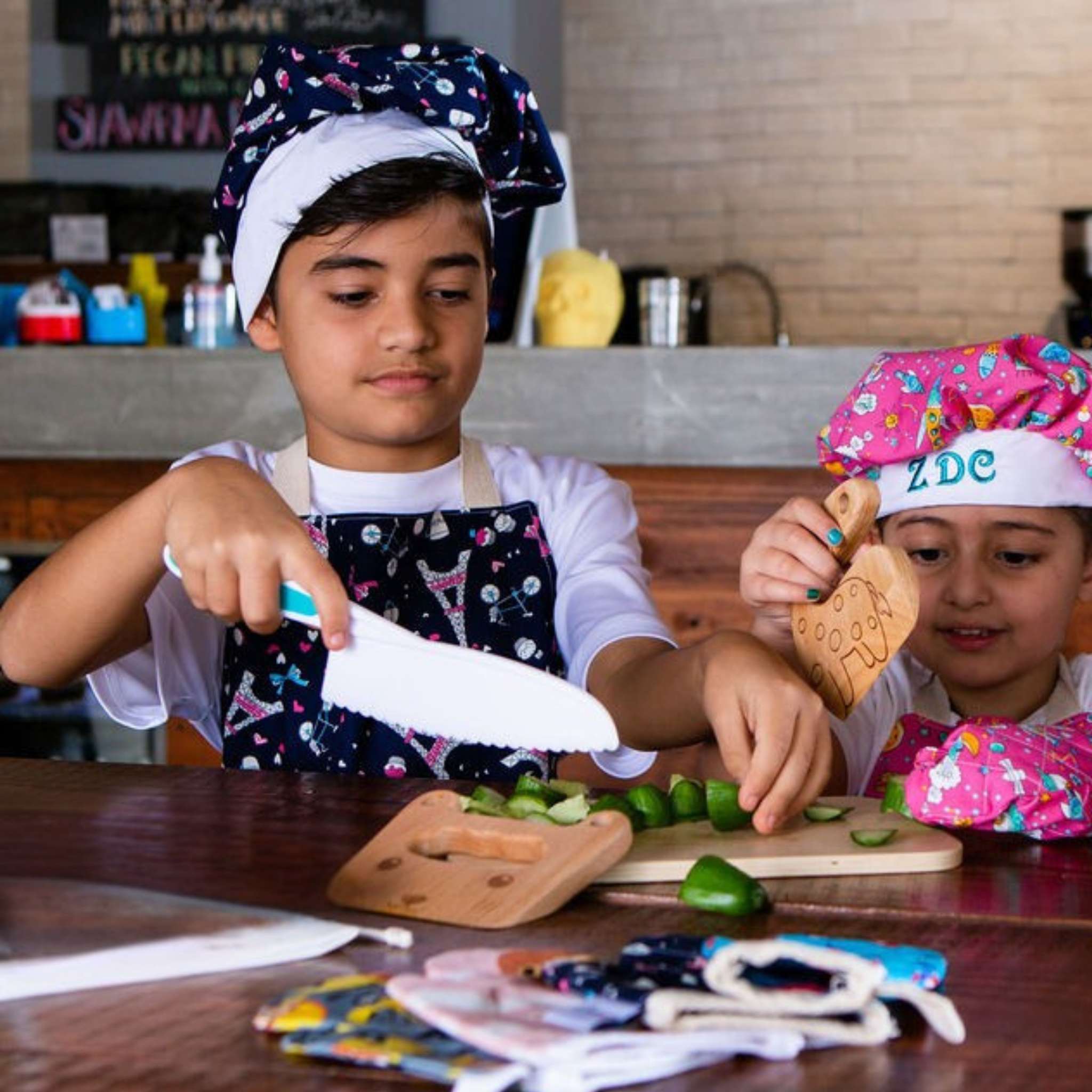 Two children in a kitchen setting, one cutting vegetables and the other holding a cookie, both wearing chef hats and aprons.
