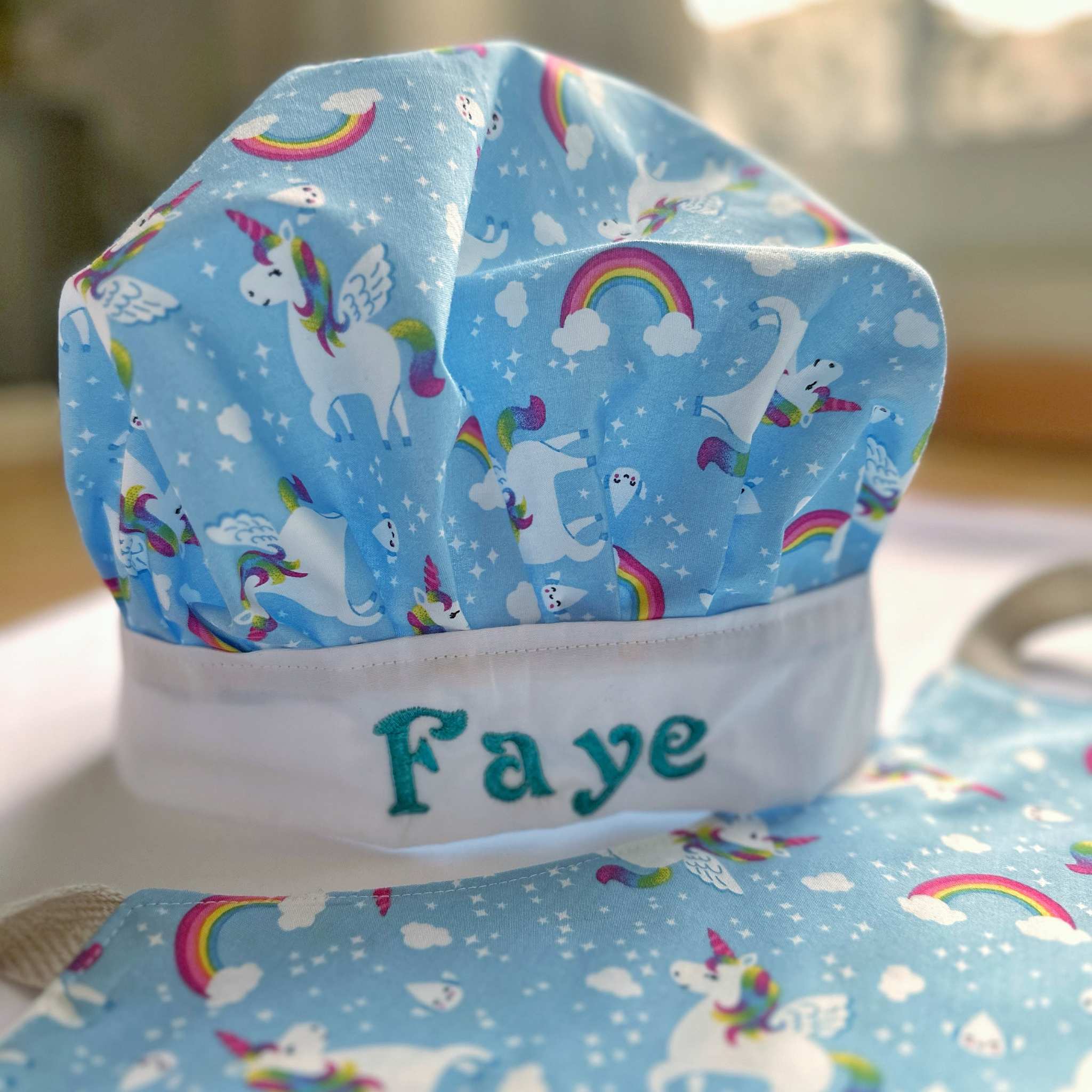 Blue chef's hat with unicorn and rainbow patterns and 'Faye' embroidered on it.