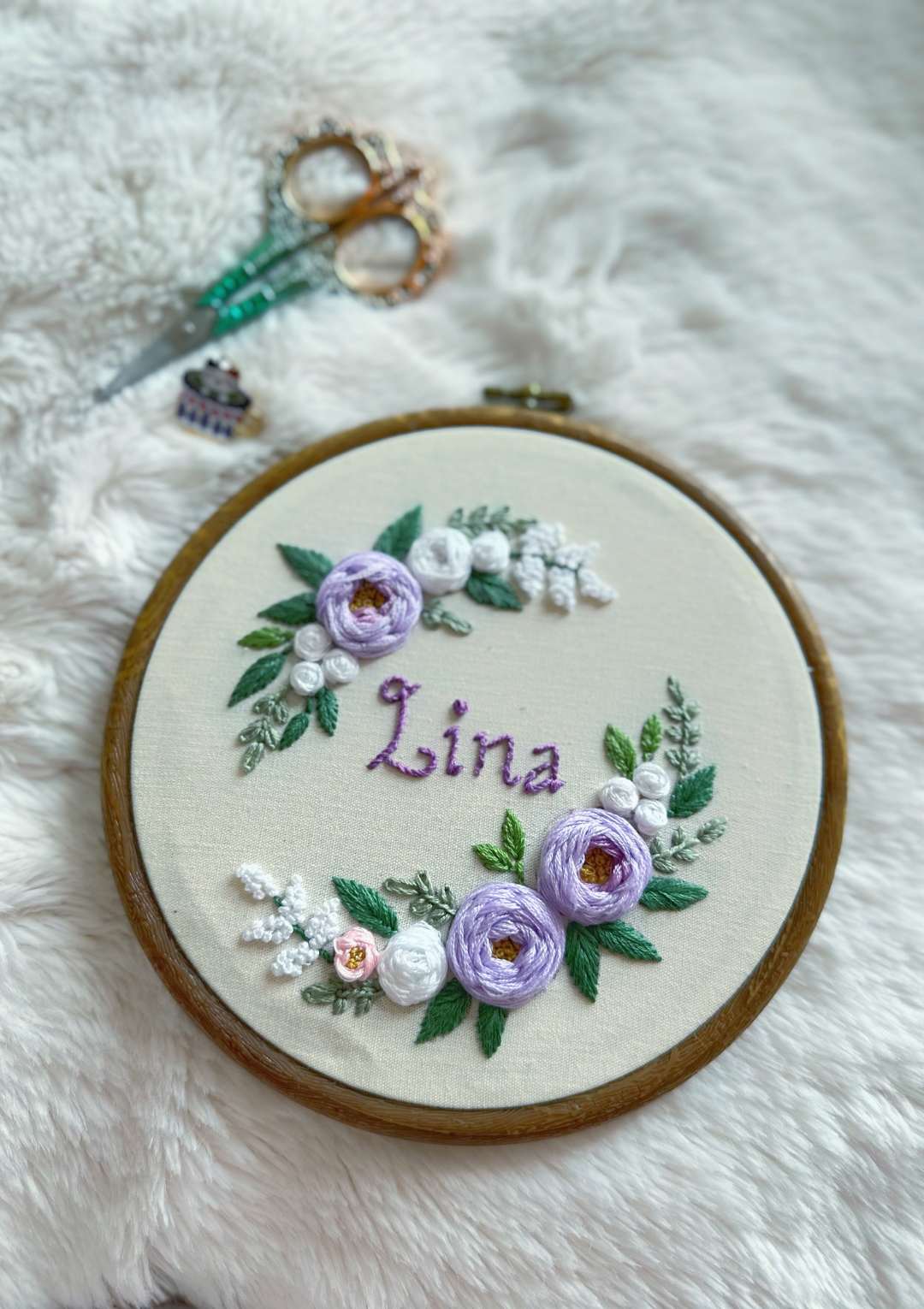 Embroidery hoop with floral design and 'Lina' text on a fluffy white surface.