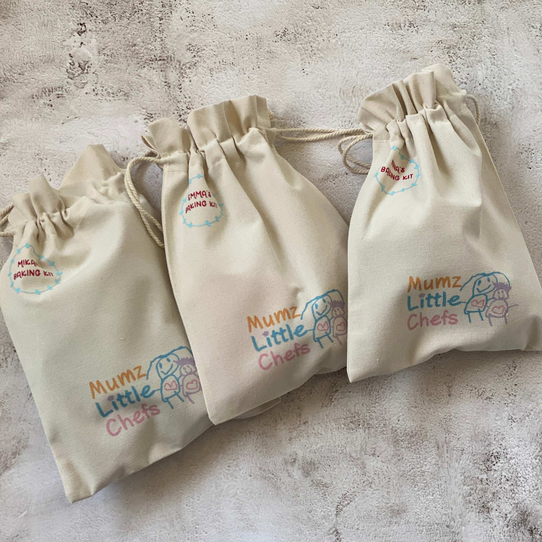 Three beige drawstring bags with 'Mumz Little Chefs' branding on a textured surface.