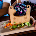 Load image into Gallery viewer, Wooden cutting board with vegetables and a child's hand, with a colorful towel on top.
