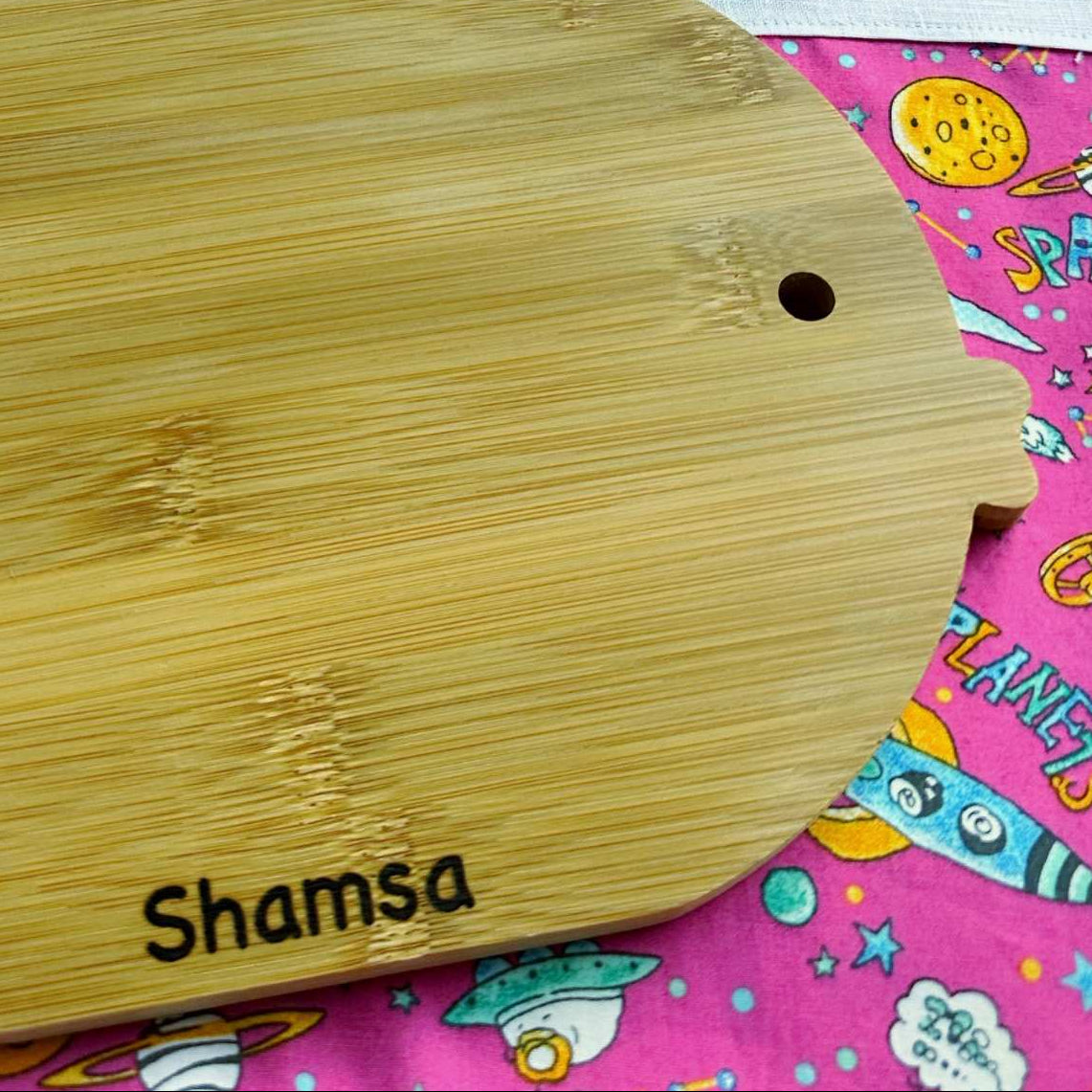 Wooden cutting board with 'Shamsa' branding on a colorful fabric background